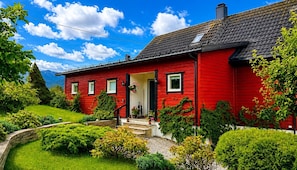 Une maison rouge avec un toit noir, entourée de verdure et sous un ciel dégagé.