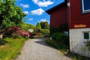 A red building with a gravel path leading to it, surrounded by lush greenery and flowering plants.