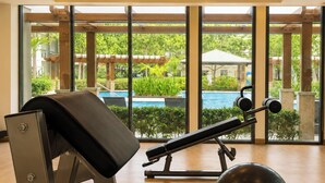 Uma academia com equipamentos de ginástica, uma janela grande e vista para uma piscina e vegetação.