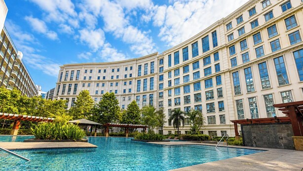 Um hotel com piscina, rodeado por vegetação e um edifício grande com várias janelas.