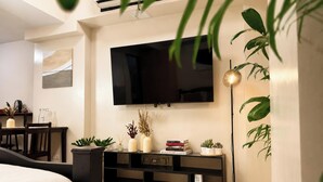 Uma sala de estar moderna com uma TV de tela plana na parede, um móvel de entretenimento em madeira escura com livros e objetos decorativos, e um abajur de chão alto.