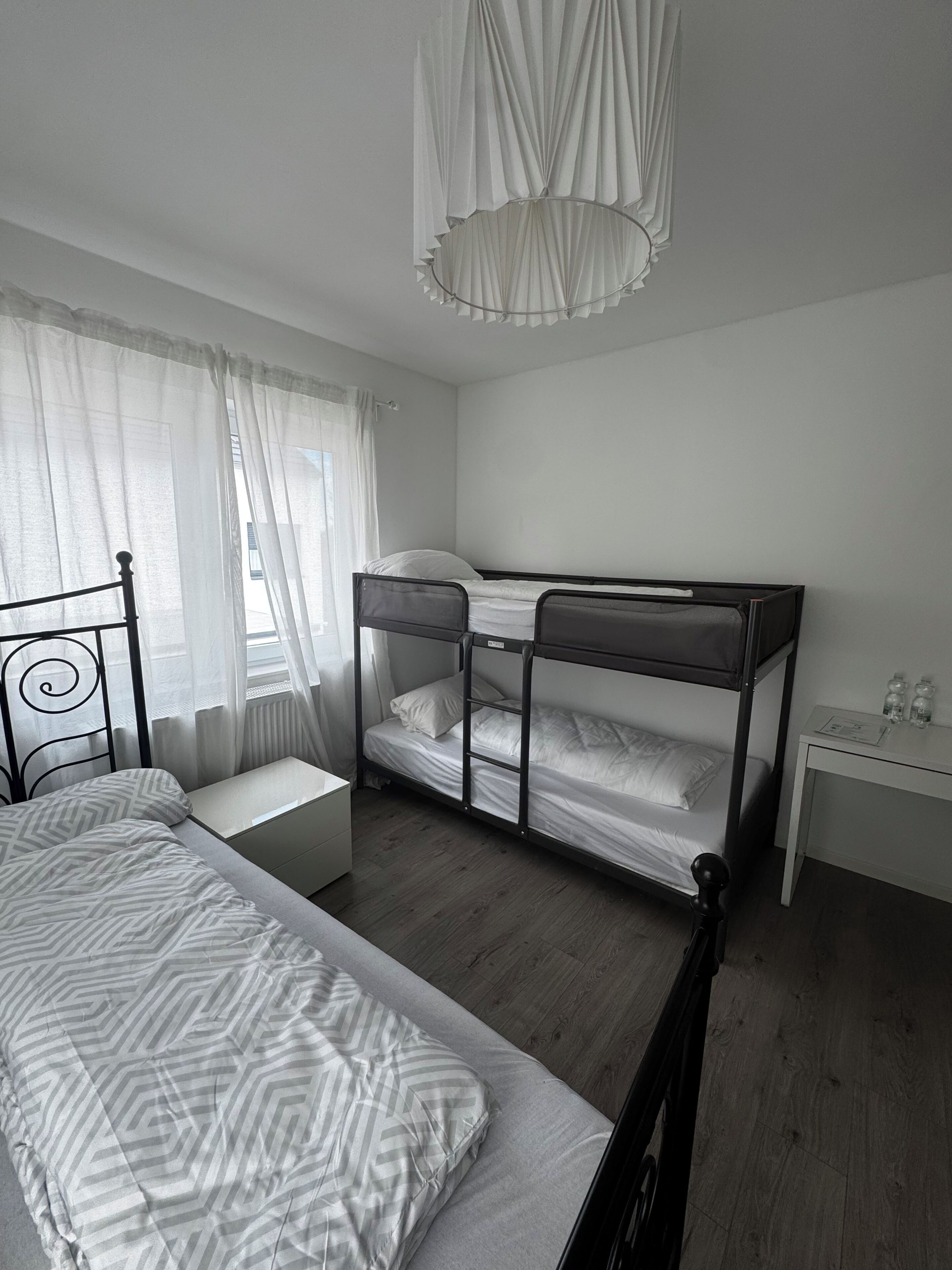 A bedroom with a bunk bed, a single bed, a bedside table, a desk, and a hanging light fixture.