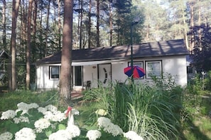 A small white house with a dark roof, surrounded by tall trees and a garden with white flowers. - Kagarsee holiday resort (Rheinsberg, OT Kagar)