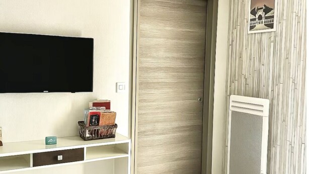 Quarto com porta de madeira, TV fixada na parede, escrivaninha com espaço de armazenamento e papel de parede estampado.