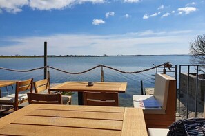 En træterrasse med borde og stole med udsigt over et roligt vandareal, et rekkerværk og en bro i det fjerne.