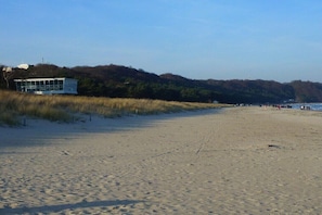 Ein Strand mit einem Gebäude links, sandigem Gelände und einer bewaldeten Hügelkette im Hintergrund.