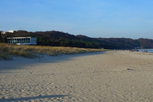 A beach with a building on the left, sandy terrain, and a forested hill in the background. - Holiday apartment in Baabe with swimming pool (Baabe)