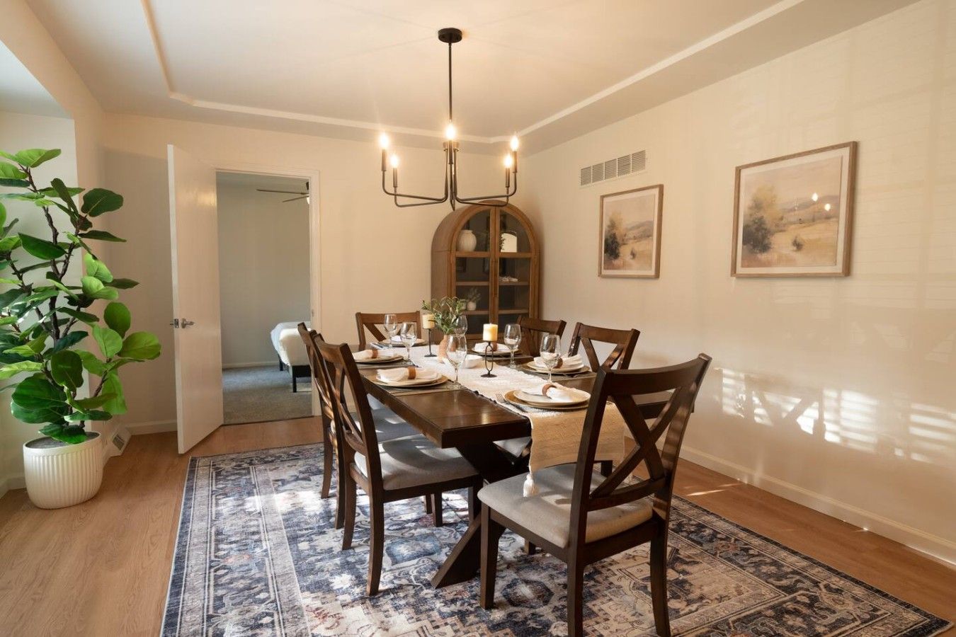 A dining room with a wooden table set for four, surrounded by chairs, a chandelier, and a large potted plant.