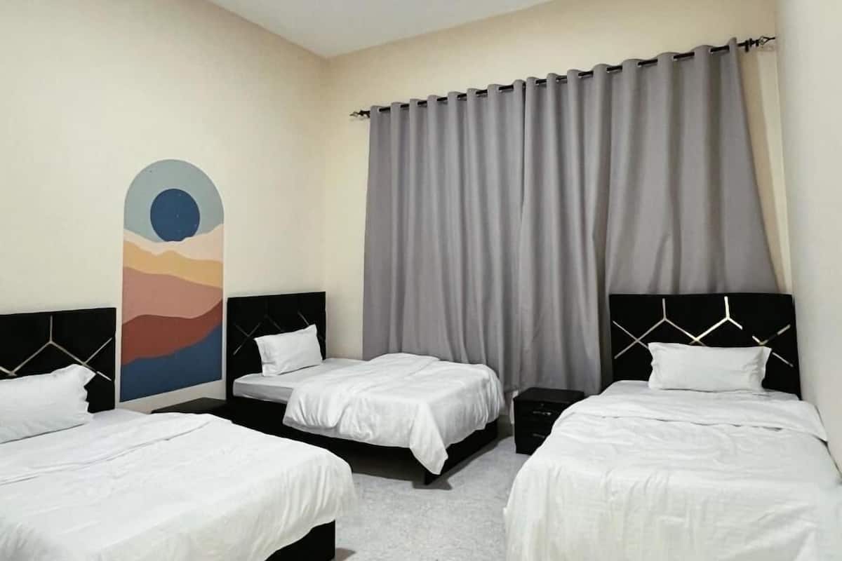A hotel room with three beds, a large window with grey curtains, and a wall painting.
