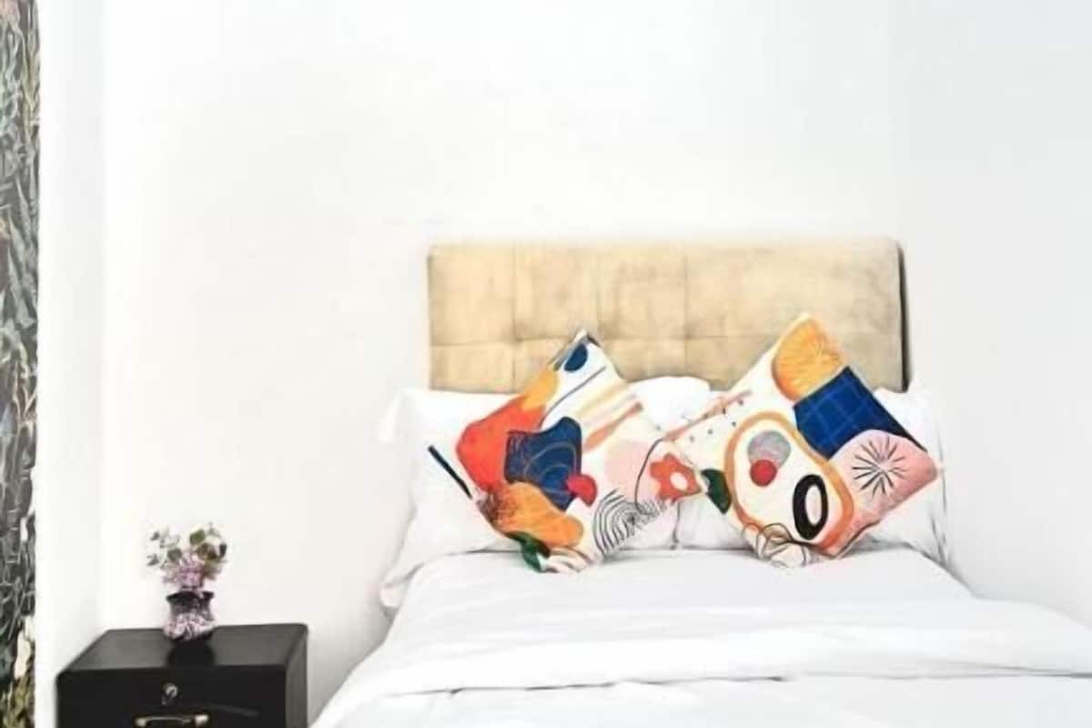 A bedroom with a bed, two pillows with a colorful design, a nightstand with a small plant, and a headboard.