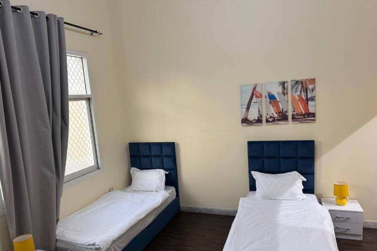 A room with two beds, a window with curtains, a wall-mounted artwork, and a bedside table with a lamp.