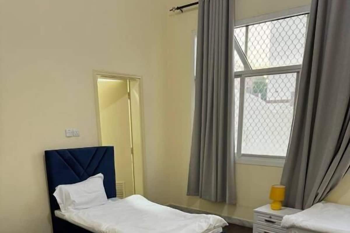 A hotel room with two single beds, a small bedside table with a lamp, a window with curtains, and a door leading to another room.