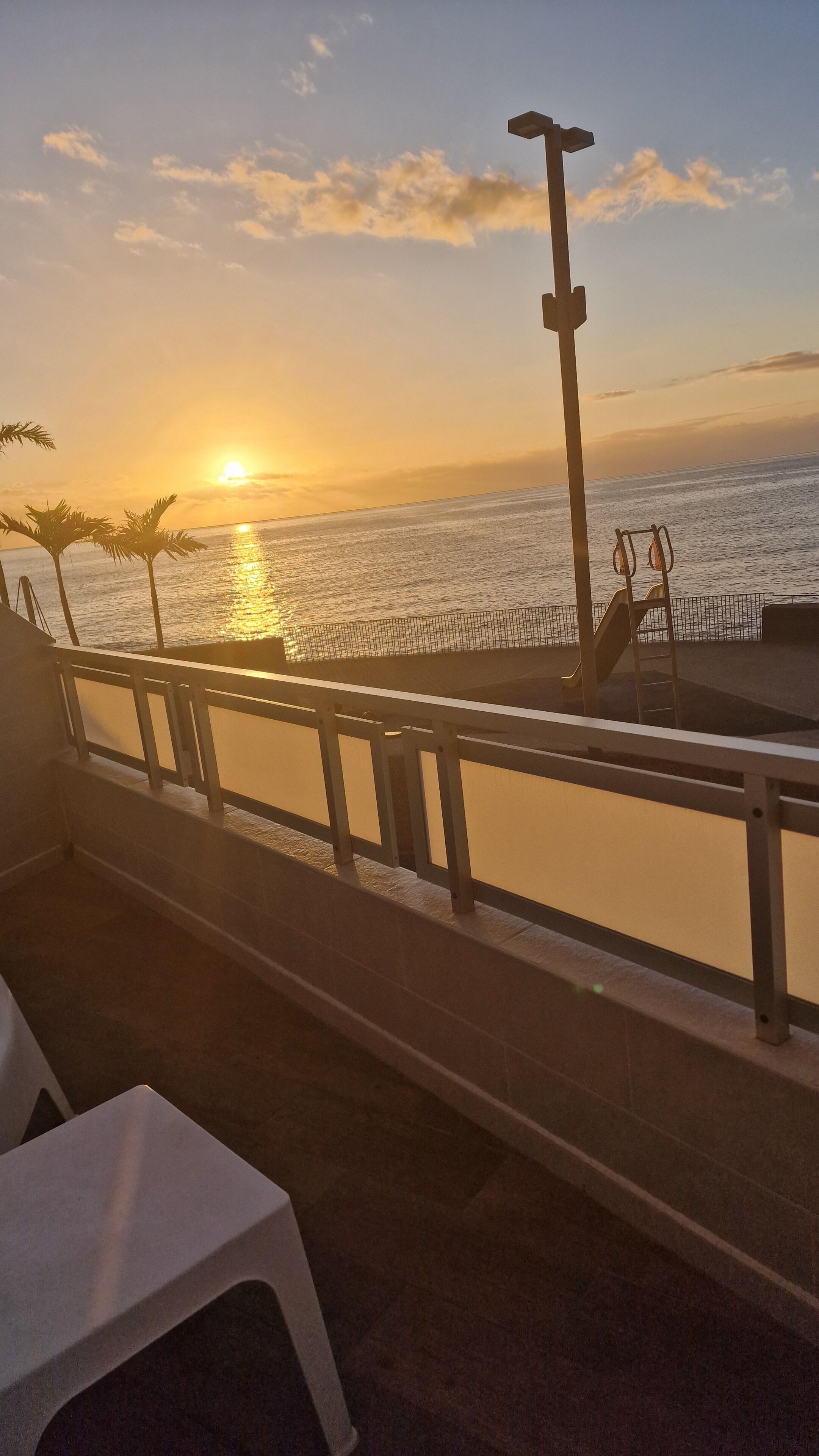 Zonsondergang zicht vanaf een balkon met een hekwerk, palmbomen en een metalen trap die naar het strand leidt.
