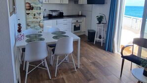 Ett kompakt kök med vita skåp, ett matbord med stolar och en utsikt över havet genom ett stort fönster.