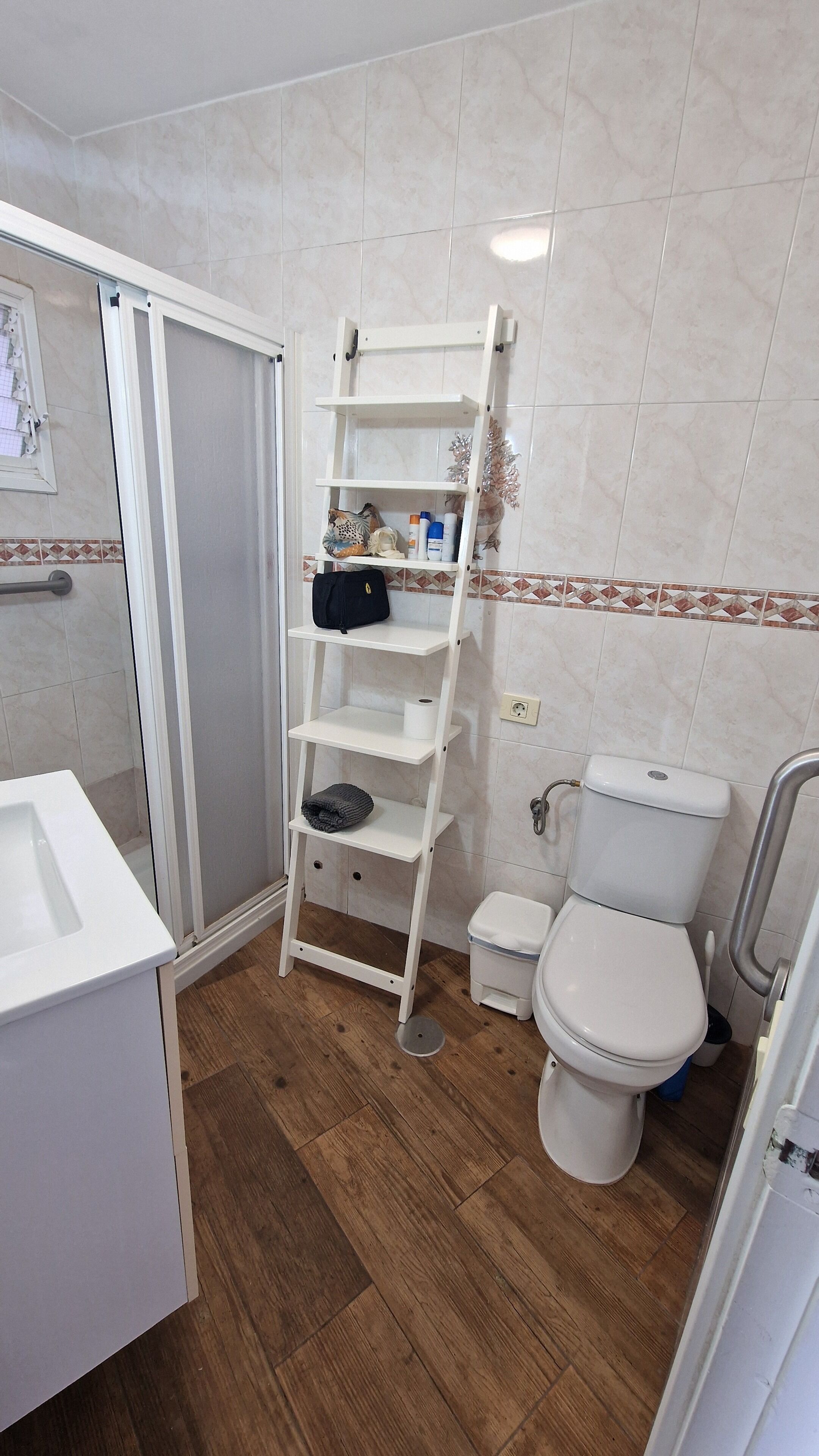 Een badkamer met een wit toilet, een witte wastafel, een houten vloer, een wit schap met toiletartikelen en een douchehoek.