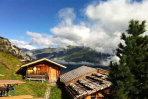 Eine Holzhütte mit geneigtem Dach, einer Bank und einem Tisch, umgeben von Bergen und Wolken.