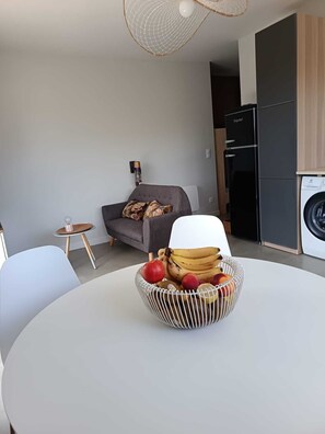 A modern living room with a round table, a sofa, a washing machine, and a compact kitchen area. - 2-room apartment for couples and families with a large, sunny terrace in an ideal location (La Teste-de-Buch)