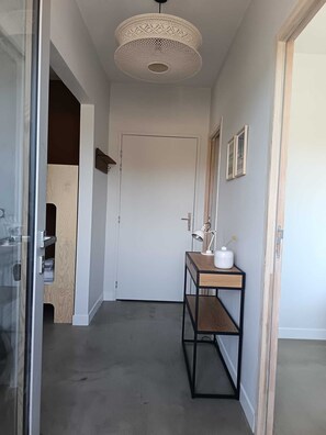 A hallway with a wooden door, a white wall, a wooden shelf with a lamp, and a ceiling light fixture. - 2-room apartment for couples and families with a large, sunny terrace in an ideal location (La Teste-de-Buch)