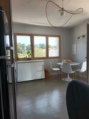 A modern kitchen with a large window, a dining area with white chairs, and a hanging light fixture. - 2-room apartment for couples and families with a large, sunny terrace in an ideal location (La Teste-de-Buch)