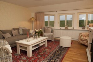 A living room with a sofa, a white coffee table, a wicker chair, a red patterned rug, and a window with a view of the outdoors. - Holiday apartment with 1 bedroom (Grünendeich)