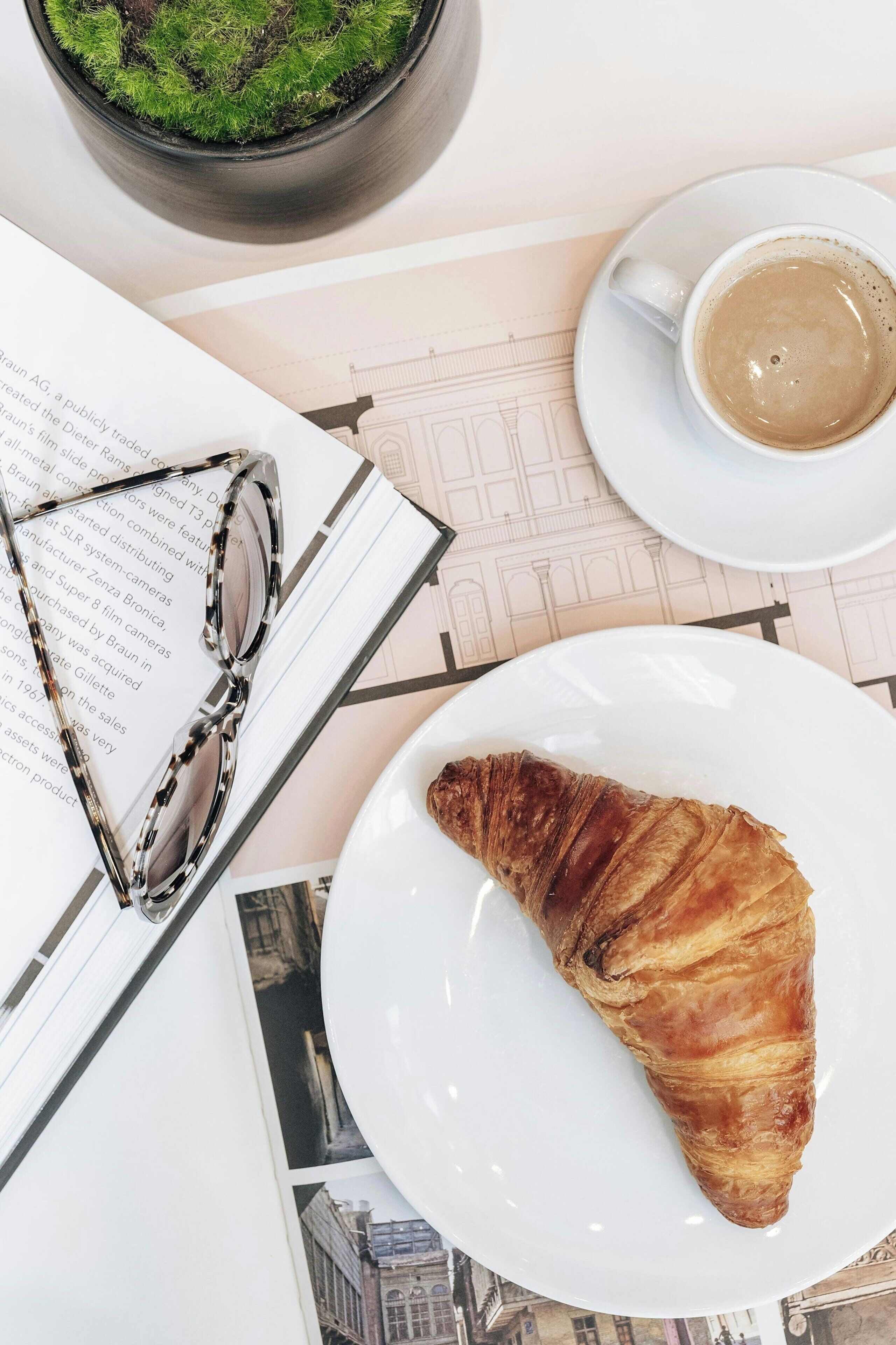A croissant on a plate, a cup of coffee, a pair of glasses, and a book with text on a table.