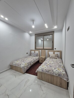 Ein Schlafzimmer mit zwei Betten, einem Fenster mit Jalousien, einem Holz-Kopfteil und einem Marmorboden.