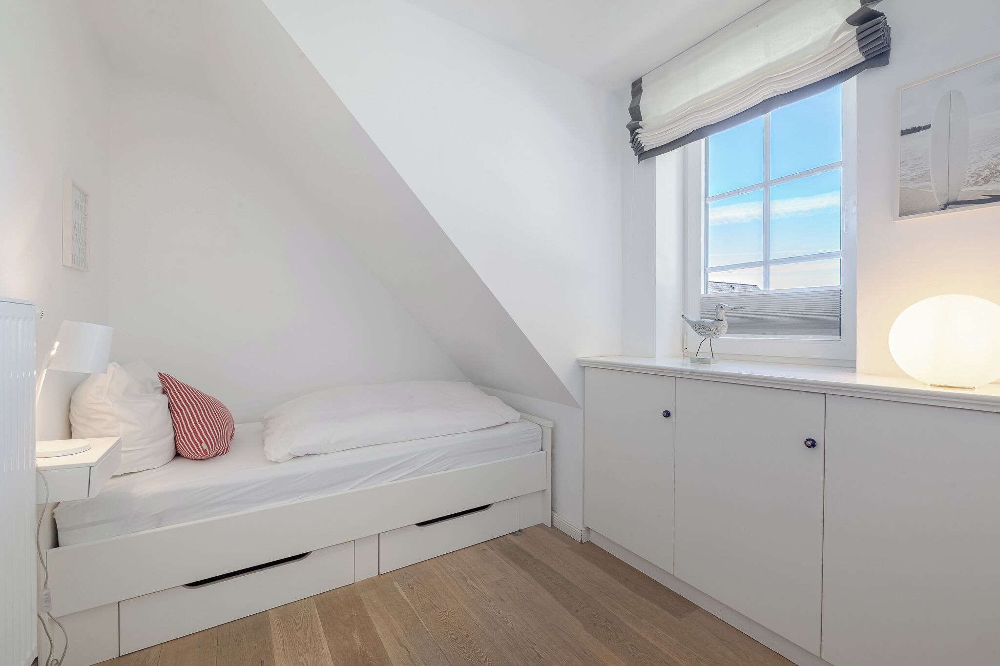 Un dormitorio estilo loft pequeño y blanco, con techo inclinado, una cama individual, una mesita de noche, una lámpara, una ventana con vistas y un armario.