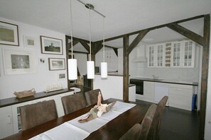 Ein Essbereich mit einem Holztisch, Stühlen und einer weißen Tischdecke. An der Wand befinden sich gerahmte Bilder, ein hängendes Licht und im Hintergrund eine Küche.