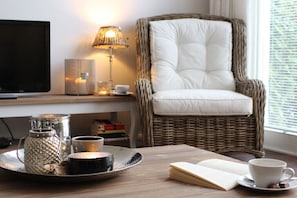 A wicker chair with a cushioned seat, a tray with a teapot, a cup, and a book, a lamp, and a television on a wooden table. - E 18 in the house with sea view (Kühlungsborn)