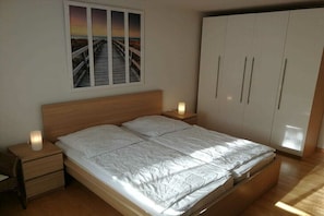 Ein Schlafzimmer mit einem hölzernen Bett, einem Nachttisch mit Lampe, einem Kleiderschrank und einem Fenster mit Sonnenuntergangsblick.