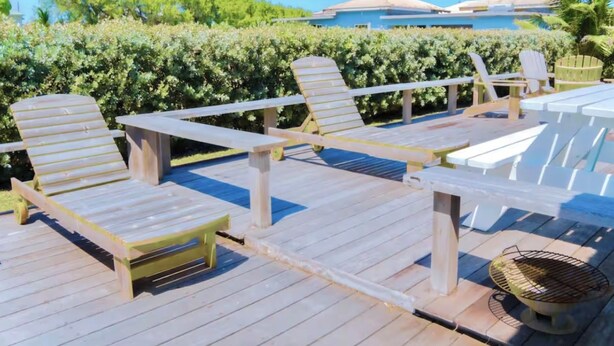 A wooden deck with lounge chairs and a grill area, overlooking a pool and a building with a blue roof. - Endless Summer - by ZenBreak (Inch Marlow)