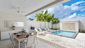 Un espace de restauration extérieur moderne, doté d’une piscine, de mobilier blanc et de palmiers.