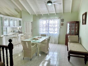 A dining area with a glass table and wicker chairs, a kitchen with white cabinets, and a wooden cabinet with glassware. - Tropical Retreat - by ZenBreak (Heywoods)