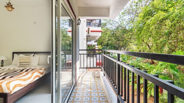 A balcony with a patterned floor, a bed with a striped blanket, and a view of greenery and a building in the background.