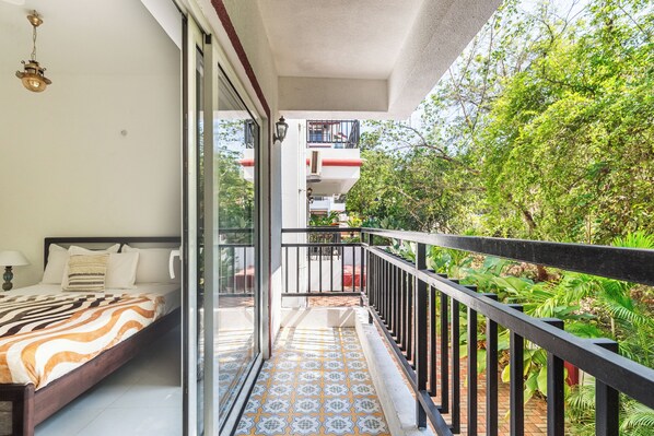 A balcony with a patterned floor, a bed with a striped blanket, and a view of greenery and a building in the background.