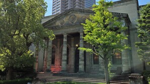 Ein historisches Gebäude mit klassischer Architektur, darunter Säulen und ein Giebel, umgeben von gepflegter Bepflanzung.
