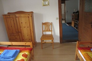 Ein Zimmer mit zwei Holzbetten, einem Holzstuhl, einem Kleiderschrank und einer Tür zu einem weiteren Raum.