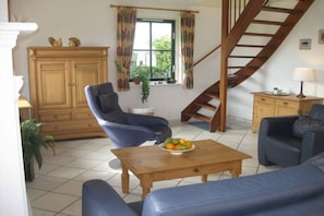 Ein Wohnzimmer mit Treppe, einem Holzschrank, zwei blauen Sessel und einem Holztisch mit Früchten sowie einem Fenster mit Vorhängen.