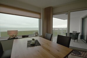 Quarto com uma janela ampla com vista para um corpo de água, uma mesa de madeira e uma varanda com cadeiras.