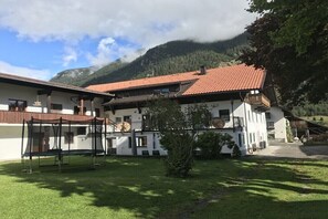 A two-story building with a red-tiled roof, a covered outdoor seating area, and a mountain backdrop. - Holiday apartment Drexel 1 (Bichlbach)