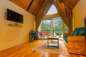 A room with a wooden ceiling, a flat-screen TV mounted on the wall, a wooden table with a rug, a sofa, and a view of greenery through large windows.