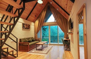 Un loft espacioso con techo alto, una escalera de madera, un sofá verde, una mesita y un balcón con vistas a árboles.