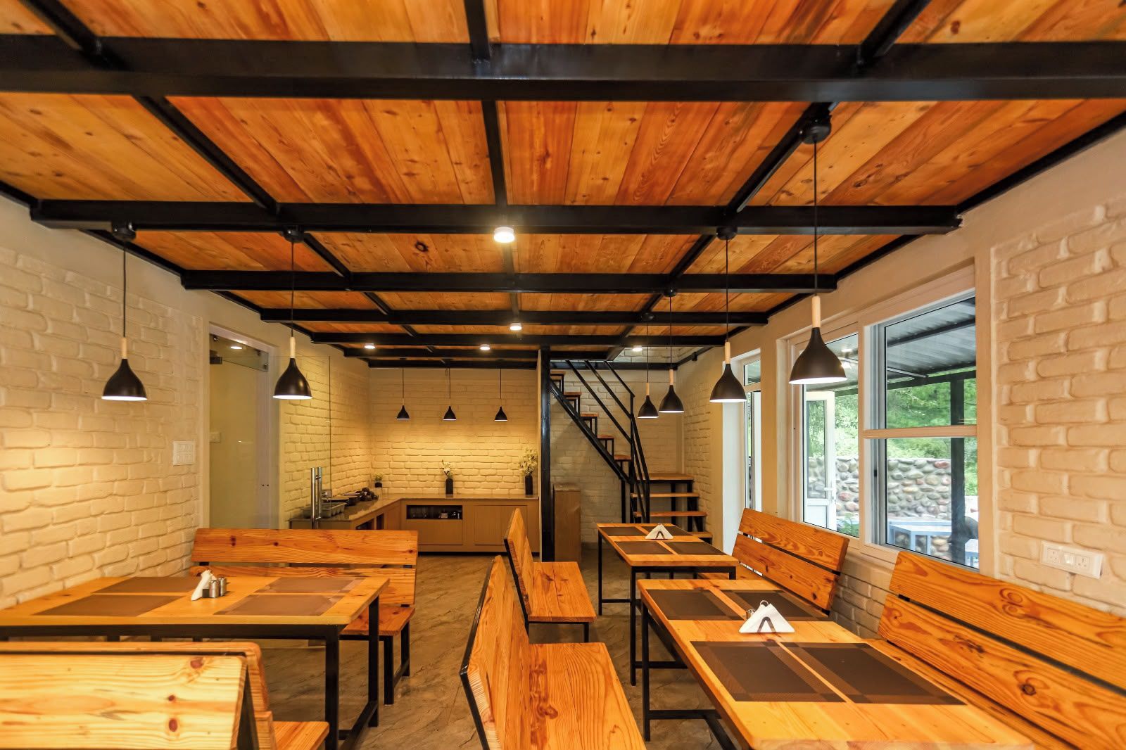 A restaurant interior with wooden tables and benches, brick walls, and a staircase leading to a kitchen area.