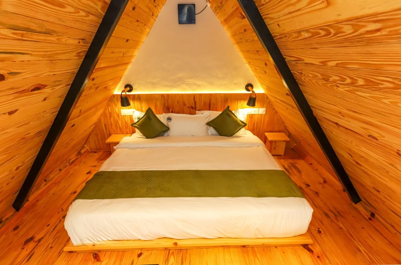 Une pièce triangulaire en bois, comprenant un lit, deux tables de chevet et des lampes fixées au mur.