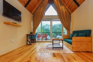 A room with a wooden ceiling, a large window with curtains, a TV mounted on the wall, a wooden sofa, a small table, and a red chair.