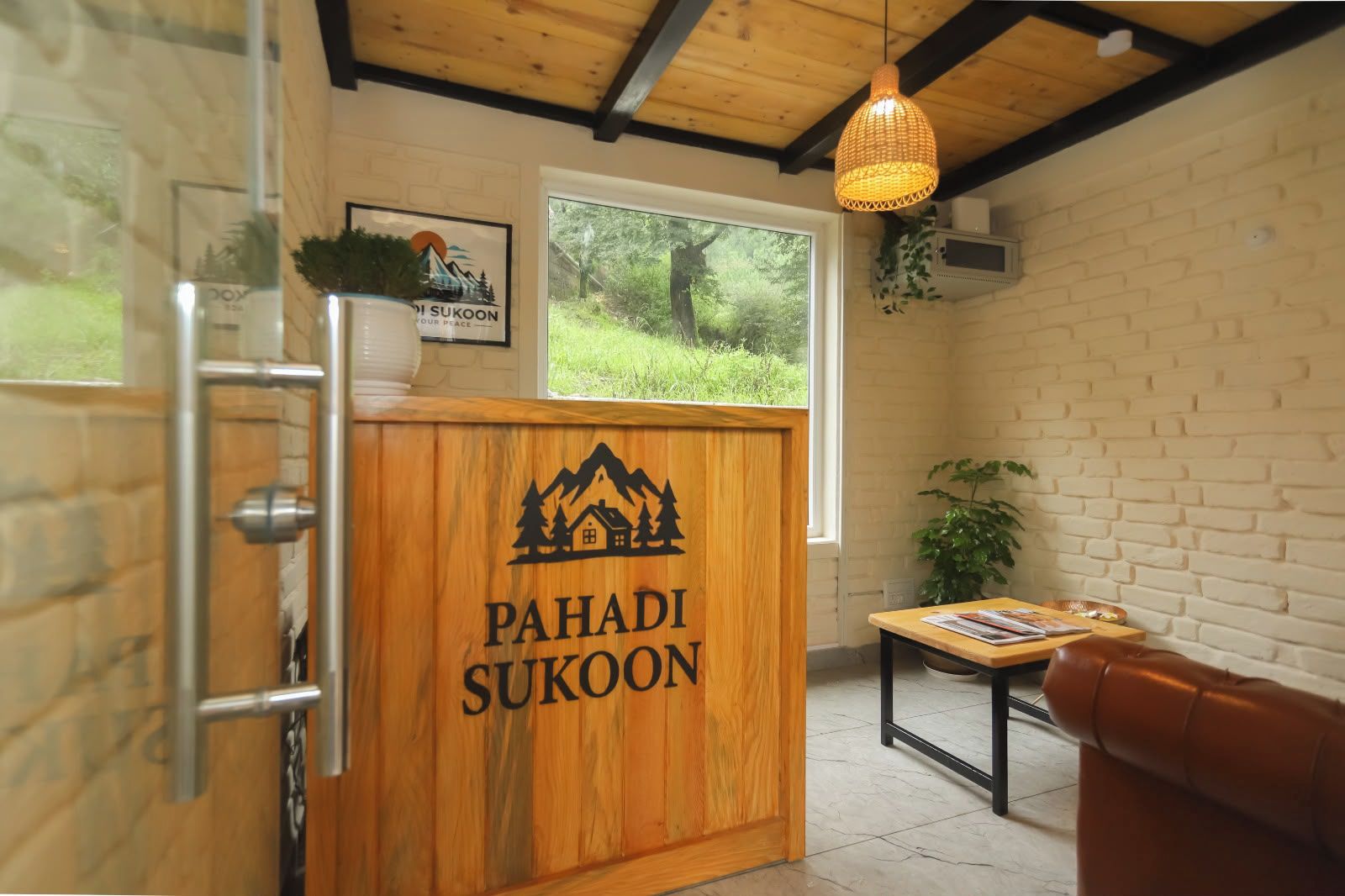 Μια ξύλινη πόρτα με την επιγραφή «PAHADI SUKOON» και ένα λογότυπο με θέμα το βουνό, ένα παράθυρο με θέα στην καταπράσ ινη φύση, ένα κρεμαστό φωτιστικό και ένα μικρό τραπέζι με διάφορα αντικείμενα.