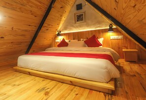 Un acogedor dormitorio en el ático con techo inclinado de madera, una cama con ropa blanca y almohadas rojas, y una mesita de noche pequeña.