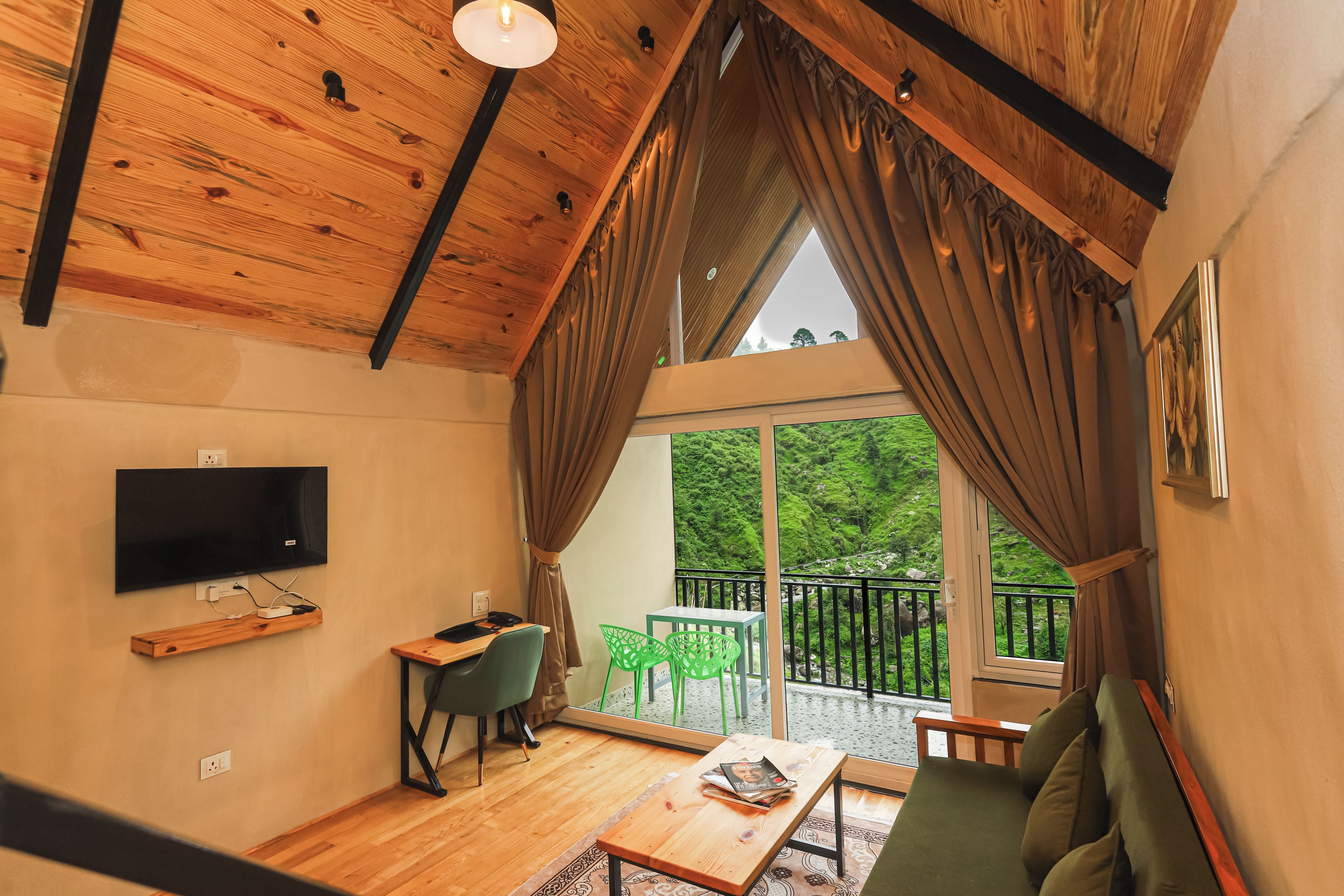 A room with a wooden ceiling, a flat-screen TV, a desk with a chair, a sofa, and a balcony with a view of greenery.