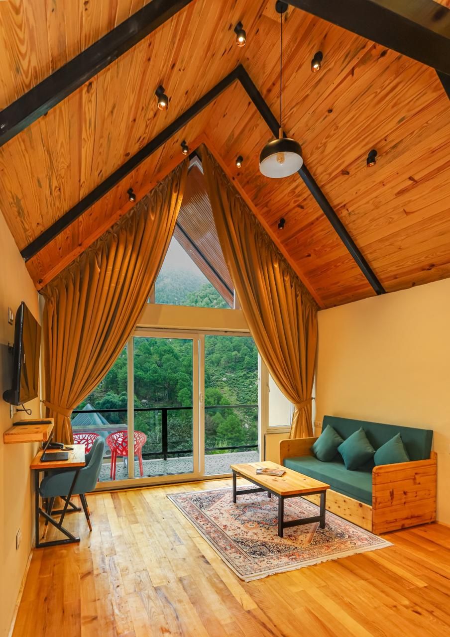 Une pièce avec un plafond en pente, un plancher en bois, un canapé vert, une table en bois et une grande fenêtre avec des rideaux.