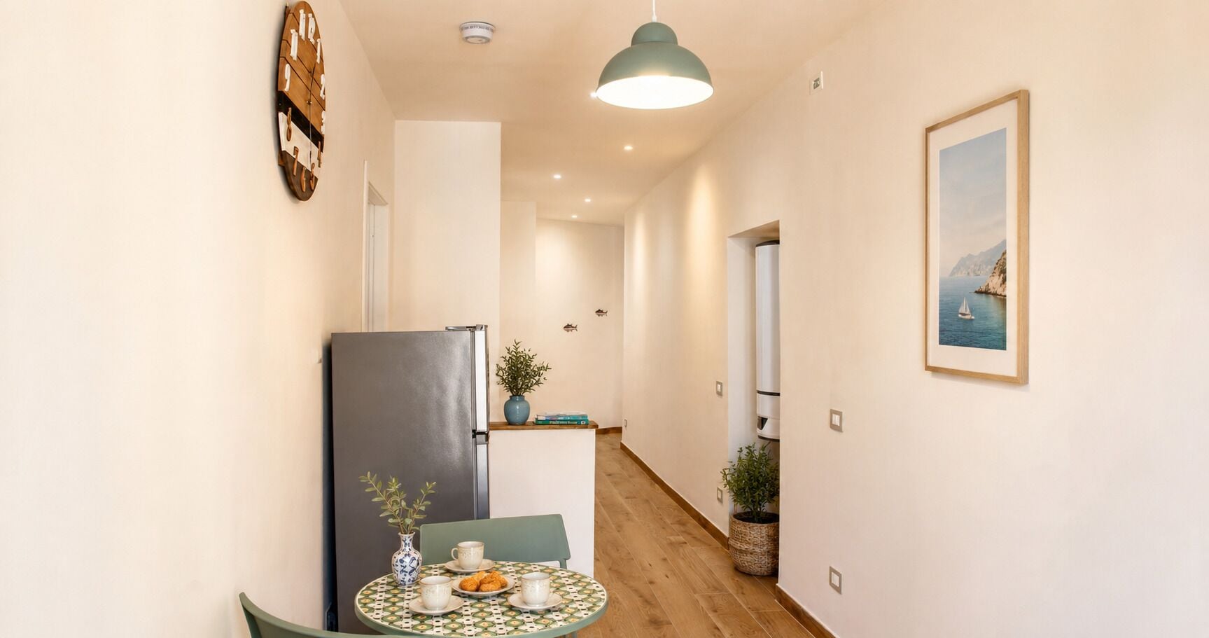 A narrow hallway with a wooden floor, a table with a vase and cups, a refrigerator, a hanging light, and a framed picture on the wall.